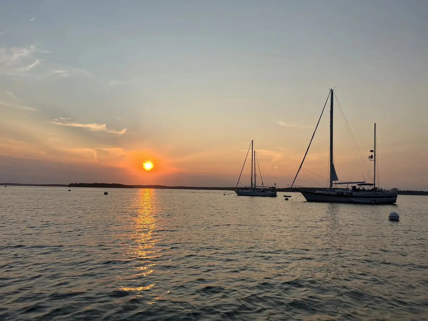 Sunset over calm water with two sailboats anchored near a distant shoreline.
